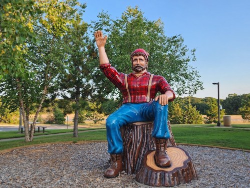 A large wooden statue of a lumberjack waving, sitting on a tree stump in a park surrounded by greenery.