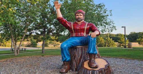 A large wooden statue of a lumberjack waving, sitting on a tree stump in a park surrounded by greenery.