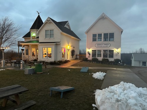 Two charming white buildings with a grassy area, one featuring a pointed roof and colorful lights, under a cloudy sky.