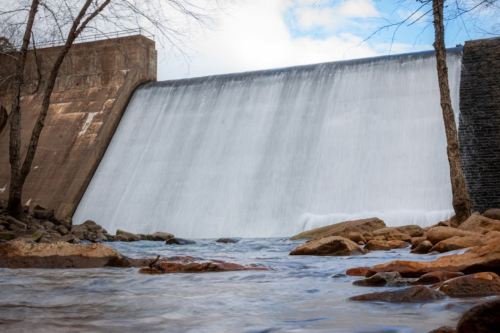 A dam with a cascading waterfall, surrounded by rocks and trees, under a partly cloudy sky.