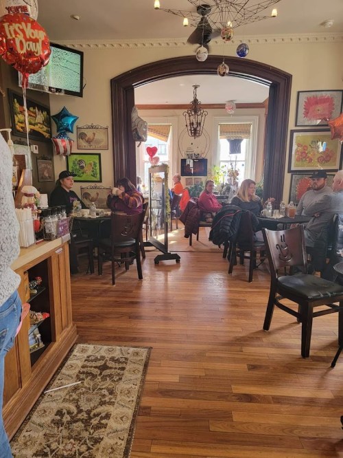 A cozy restaurant interior with wooden floors, colorful artwork, and patrons enjoying meals at tables. Balloons decorate the space.