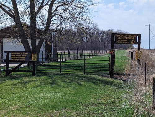 Entrance to Woodman Hollow State Preserve, featuring a wooden gate and signs, surrounded by green grass and trees.