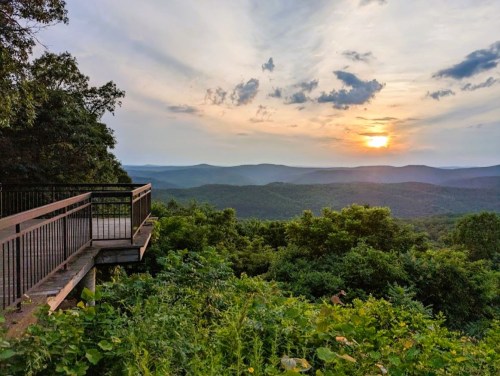 A scenic overlook with a wooden deck, surrounded by lush greenery, showcasing a sunset over rolling hills.