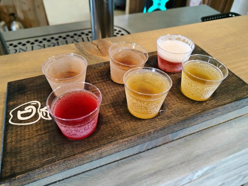 A wooden flight board with six small cups of colorful beverages, including pink, red, and golden drinks.