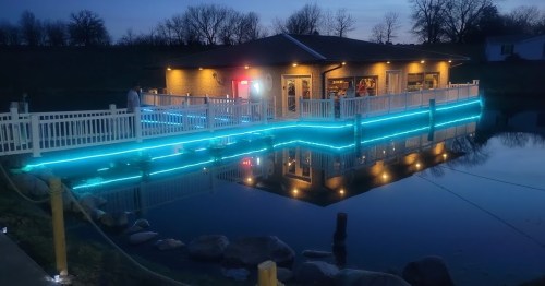 A lit building by a calm water body at dusk, with blue lights outlining the deck and reflections in the water.