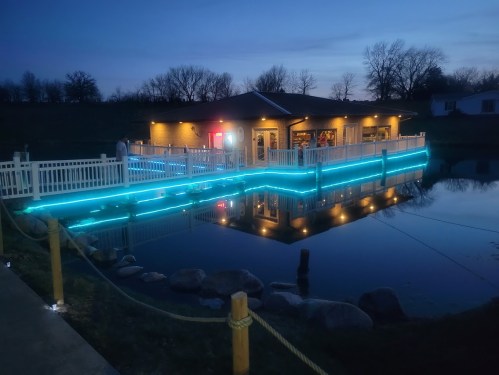 A cozy building with blue lights reflecting on a calm pond at dusk, surrounded by trees and a white fence.