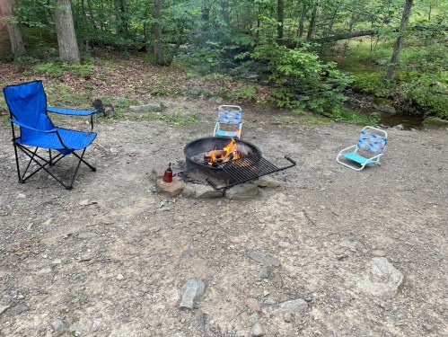 A campfire surrounded by rocks, with blue folding chairs nearby, set in a wooded area.