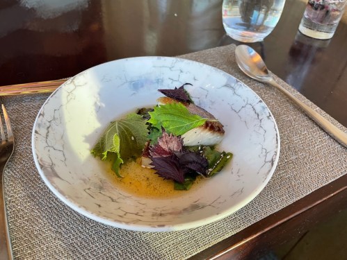 A beautifully plated dish featuring fish, fresh greens, and herbs in a light broth, served in a decorative bowl.