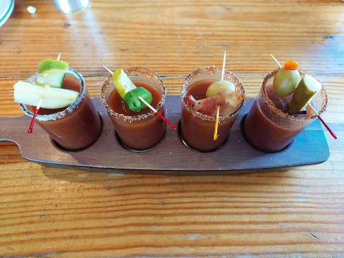 Four shot glasses of Bloody Marys garnished with celery, lime, pickles, and olives, served on a wooden tray.