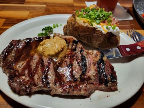 A grilled steak topped with butter, served with a baked potato loaded with toppings on a white plate.