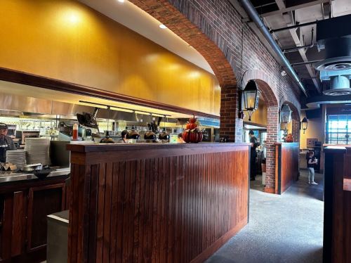 Interior of a restaurant featuring a wooden counter, brick arches, and a bustling kitchen in the background.