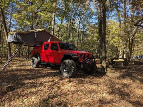 A red Jeep with a rooftop tent parked in a wooded area, surrounded by autumn foliage and picnic tables.