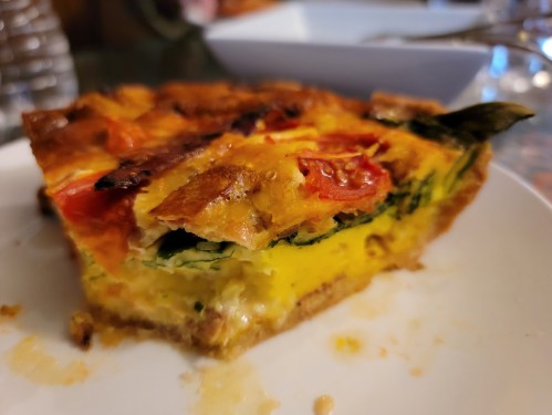 A slice of quiche with tomatoes and greens on a white plate, showcasing a golden crust and creamy filling.