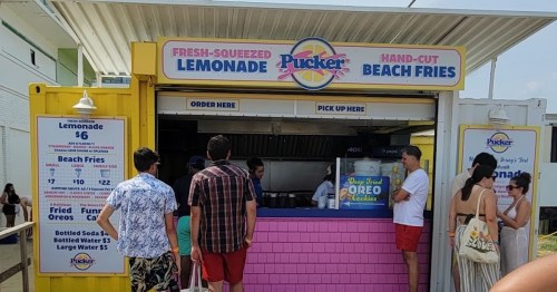 A colorful food stand selling lemonade and beach fries, with customers waiting in line on a sunny day.
