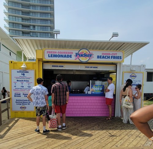 A colorful beach stand selling lemonade and fries, with people ordering and enjoying the sunny day.