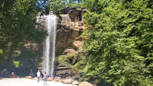 A waterfall cascades down a rocky cliff surrounded by lush green trees, with people enjoying the scenery below.