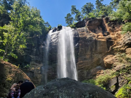 A waterfall cascades down a rocky cliff surrounded by lush greenery under a clear blue sky.