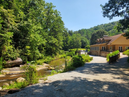 A serene landscape featuring a winding path beside a creek, surrounded by lush greenery and a cozy building.