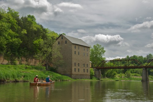Two people in a canoe paddle on a calm river near a historic stone building and a bridge under a cloudy sky.