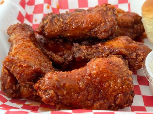 A close-up of crispy, sauced chicken wings served in a red and white checkered basket, alongside a bun and dipping sauce.