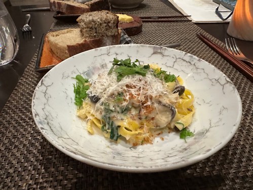 A plate of fettuccine pasta topped with herbs, cheese, and mushrooms, served with slices of bread in the background.