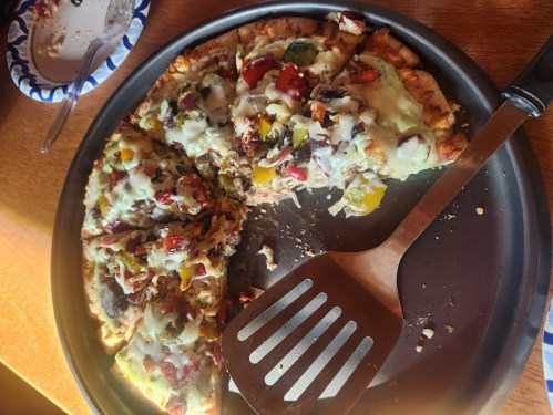 A partially eaten pizza with colorful toppings on a round tray, accompanied by a spatula and a paper plate.