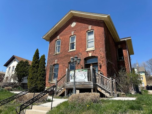 A red brick building with large windows and a wooden staircase, surrounded by grass and trees under a clear blue sky.