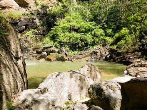 A serene natural scene featuring a calm green pool surrounded by large rocks and lush greenery.