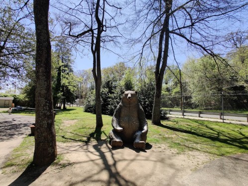 A large bear statue sits on a grassy area surrounded by trees under a clear blue sky.