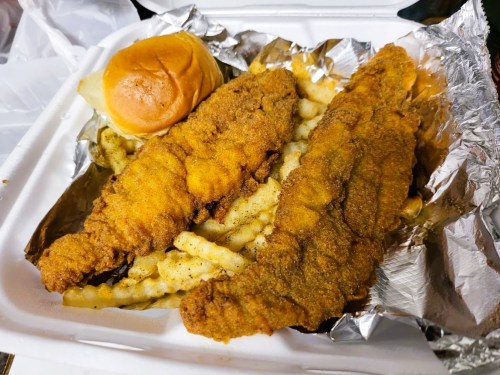 A takeout container with two fried fish fillets, crinkle-cut fries, and a soft bun.