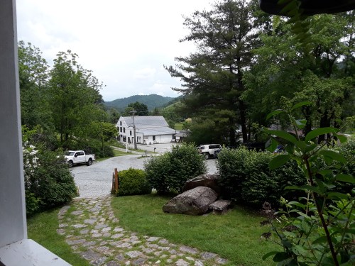 A scenic view from a porch, featuring a gravel path, greenery, and a distant white building surrounded by mountains.