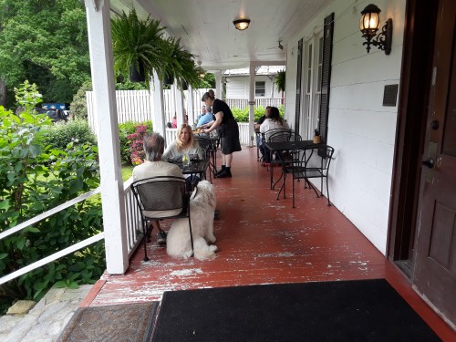 A porch with tables and chairs; people dining and a dog sitting beside a woman, surrounded by greenery.