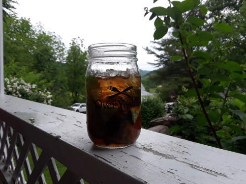 A mason jar filled with iced tea and mint sits on a porch railing, with greenery and a scenic view in the background.