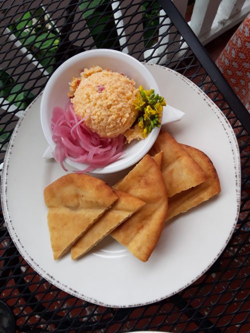 A plate with crispy flatbreads, pickled onions, and a bowl of colorful cheese spread garnished with a flower.