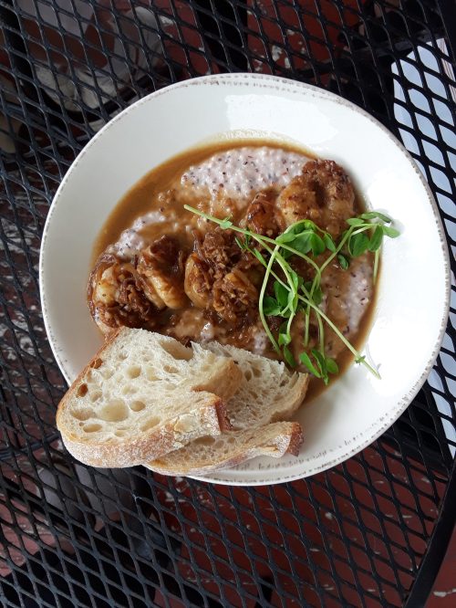 A bowl of shrimp and grits topped with microgreens, served with a slice of crusty bread on a black table.