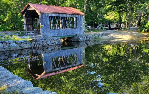 A wooden covered bridge with a red roof reflects in calm water, surrounded by trees and a peaceful landscape.