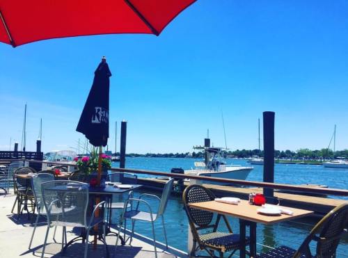 Outdoor dining area by the water, featuring tables under umbrellas and a view of boats and a clear blue sky.