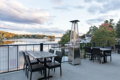 Outdoor dining area with tables overlooking a calm lake and trees, under a cloudy sky.
