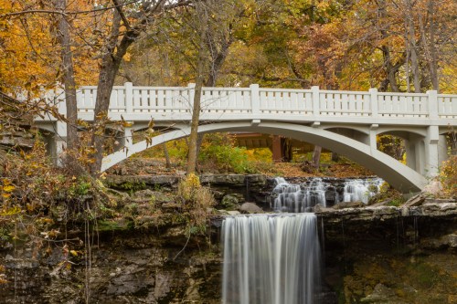 A white bridge arches over a waterfall surrounded by autumn foliage in a serene natural setting.