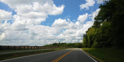 A winding road bordered by trees under a blue sky filled with fluffy white clouds.