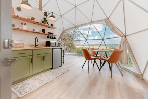 Interior of a modern geodesic dome with a kitchenette, dining table, and large windows showcasing a scenic view.