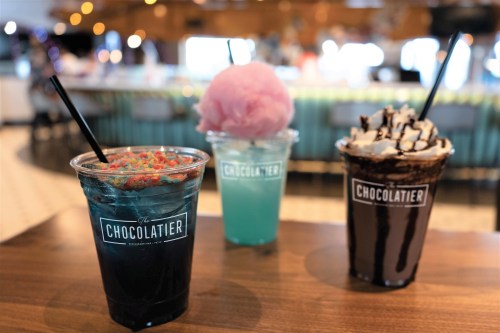 Three colorful drinks on a wooden table: a blue drink with candy, a pink cotton candy topped drink, and a chocolate drink with whipped cream.