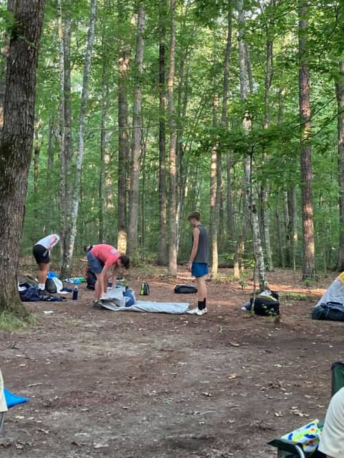 Two people set up a tent in a wooded area, surrounded by trees and camping gear.