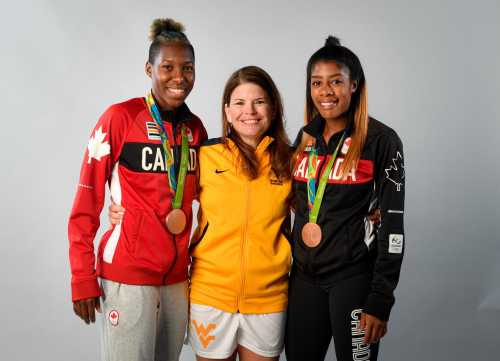 Three women pose together, two wearing Canada jackets with medals, and one in a yellow shirt, smiling against a gray background.