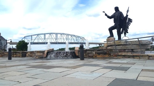 A statue of a Native American figure stands near a fountain, with a bridge visible in the background under a cloudy sky.