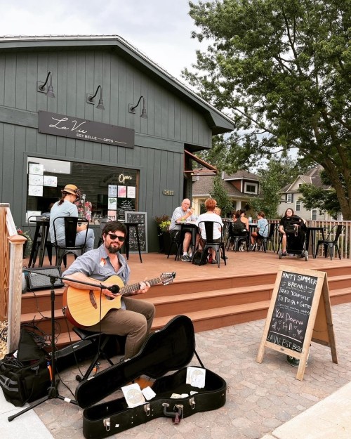 A musician plays guitar outside a café, with patrons enjoying their meals on a sunny day. A chalkboard sign is nearby.
