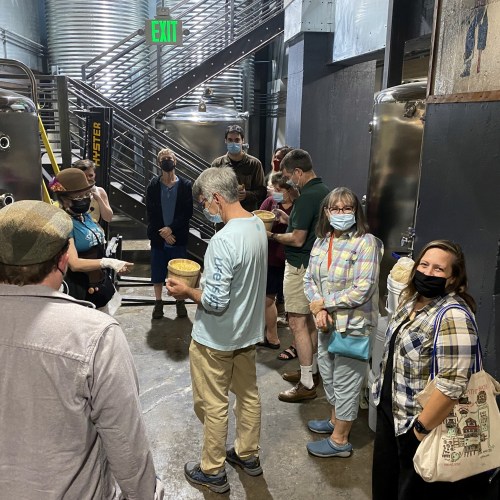A group of people wearing masks gathers in a brewery, some holding cups, while a staff member speaks to them.