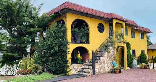 A vibrant yellow two-story house with stone accents, surrounded by greenery and colorful flowers.