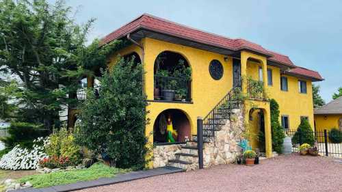 A vibrant yellow house with stone accents, surrounded by greenery and colorful flowers, featuring arched balconies.