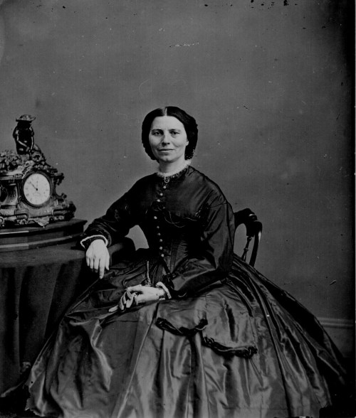 A woman in a vintage dress sits beside an ornate clock on a table, posing for a formal portrait.